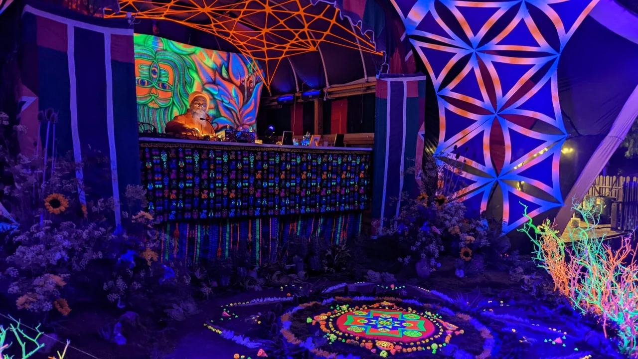 Goa Gil playing at the decks under UV light, with a glowing mandala and flowers in front of the booth